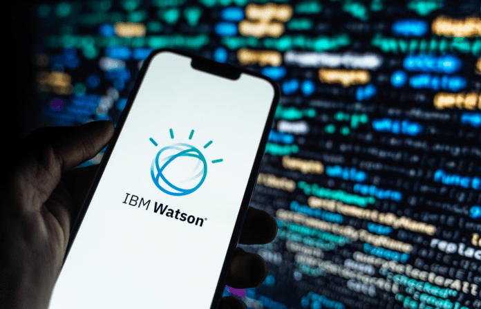 Hand holding a mobile phone with the IBM Watson logo on display on a background of program codes.